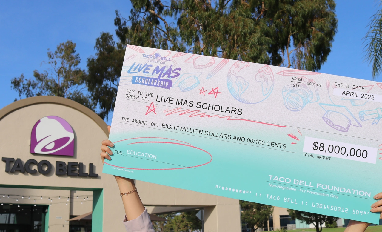 A large check in front of a Taco Bell reads " Pay to the order of : Live Más Scholarships"