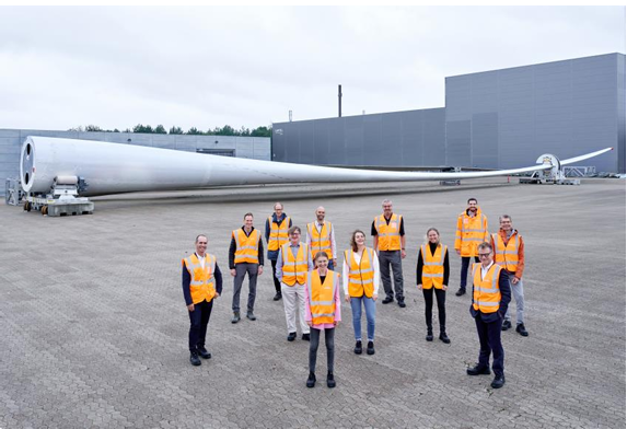 A group posed standing outside in front of a huge wind turbine blade.