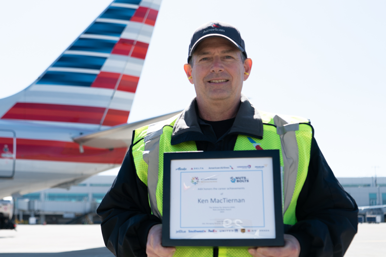 Ken MacTiernan holding a plaque outside, in front of an airplane. 