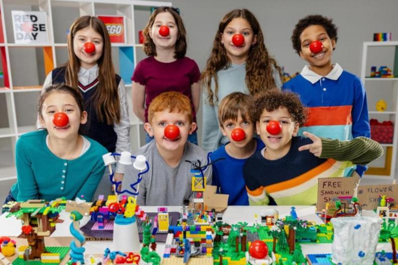 Children wearing red noses behind a table with lego models on  