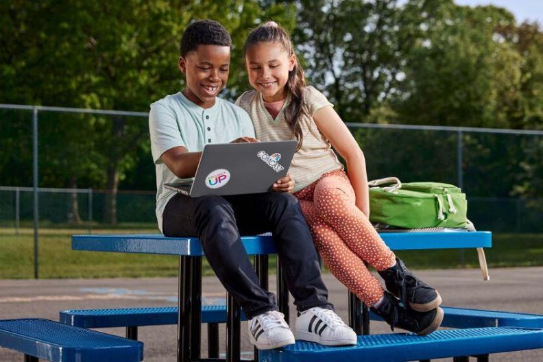 Two smiling children looking at an open laptop while sitting on an outdoor table. 