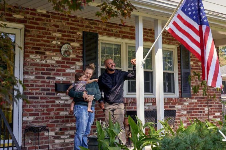Two adults and a small child standing on a porch. An american flag in a post on the side pillar. 