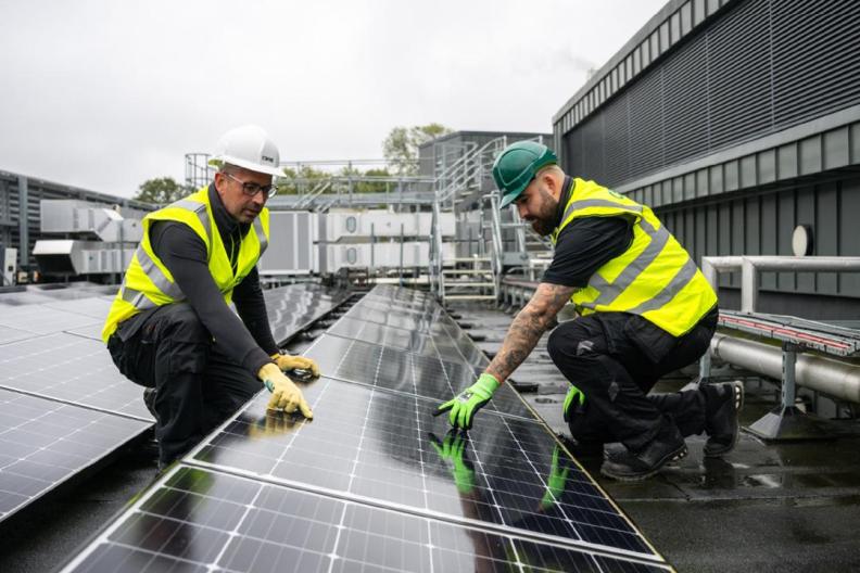 Two people in high-vis vests and hard hats pointing to a solar panel in a row of others outside. 