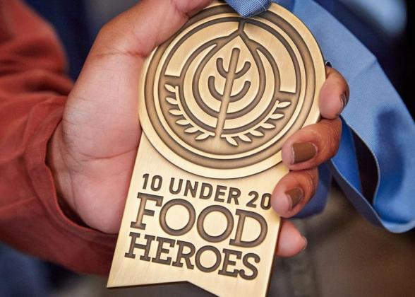 A hand holding a medal &quot;10 Under 20 Food Heroes&quot;.