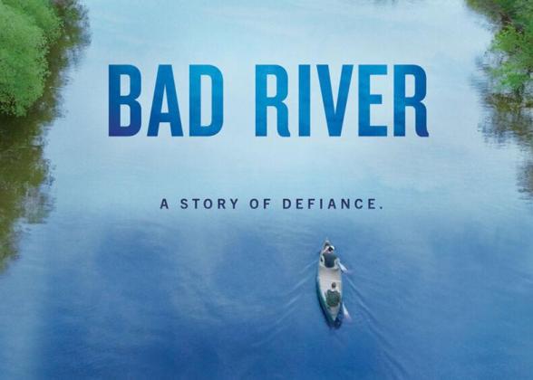 Two people in a canoe on a tree-lined river. &quot;Bad River. A story of defiance.&quot;
