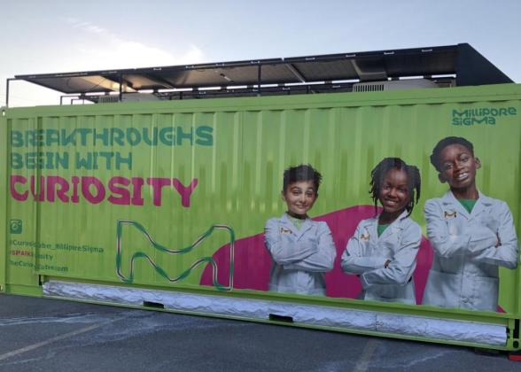 A green shipping container turned mobile science lab features three young scientists and text reading, &quot;Breakthroughs Begin With Curiosity.&quot;