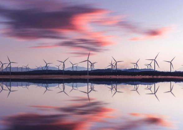 Wind turbines behind a body of water. A setting sun behind them.