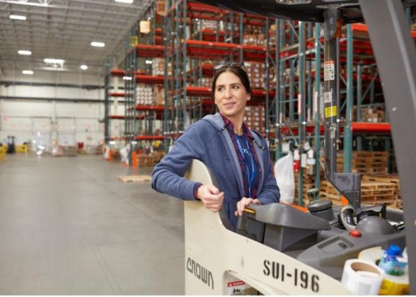 A woman in a warehouse stands in the operators seat of a forklift. She looks over her right shoulder and smiles with floor to ceiling shelves stacked with boxes behind her.