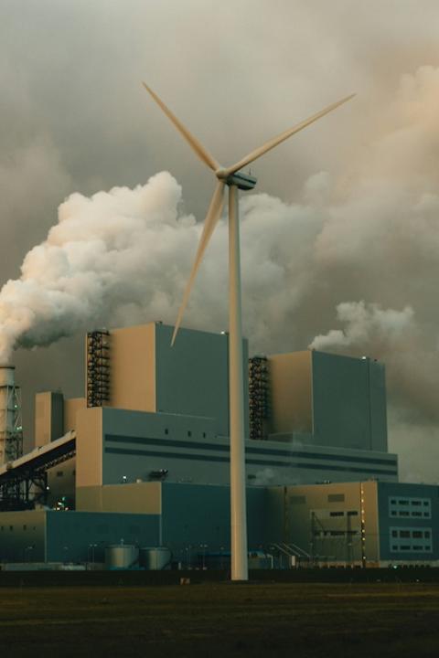 A wind turbine in front of a factory with smoke billowing out of it. 