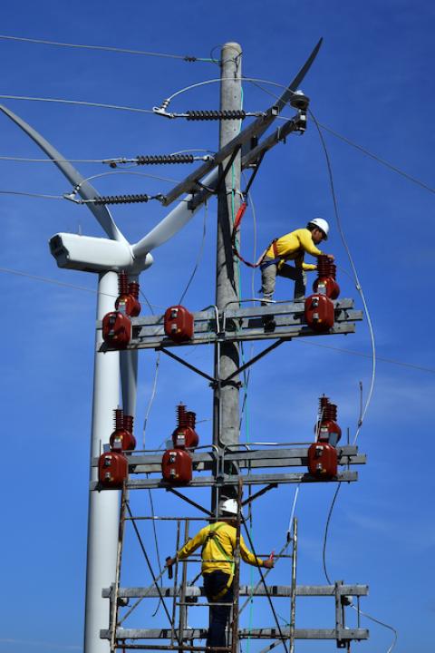 Technicians repairing an electrical pole in front of a wind turbine.