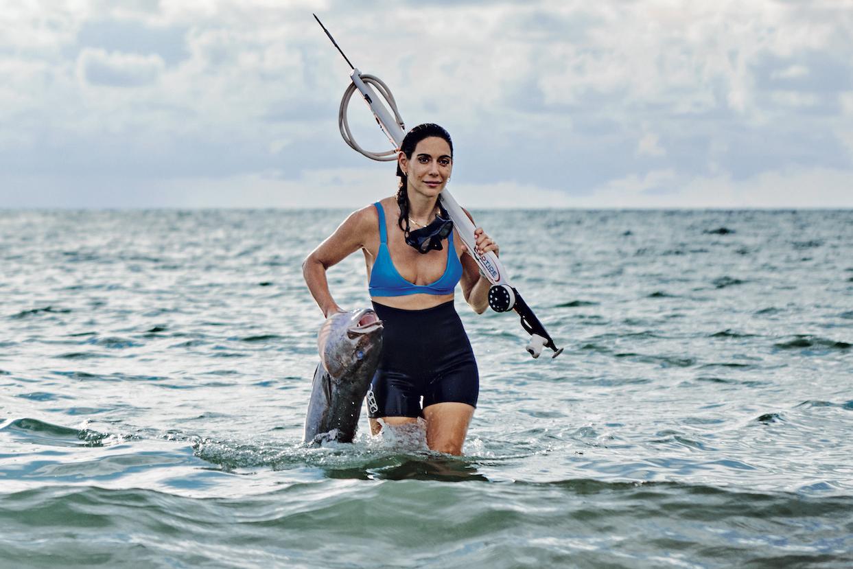 Valentine Thomas, professional spearfisherwoman and sustainable seafood advocate, emerges from the water with a fish and fishing spear.