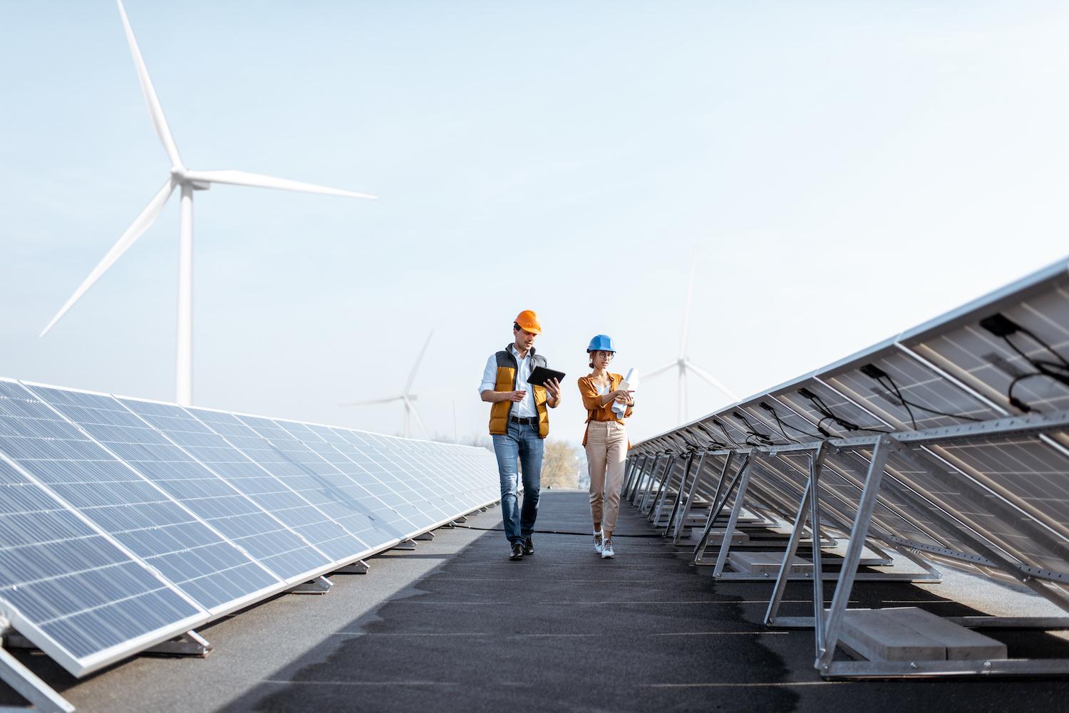Workers walking through a solar power plant with wind turbines in the background - Achieving the SDGs with renewable energy