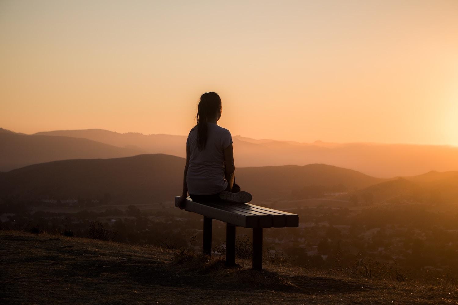 woman sitting meditating on park bench with mountain view at sunset