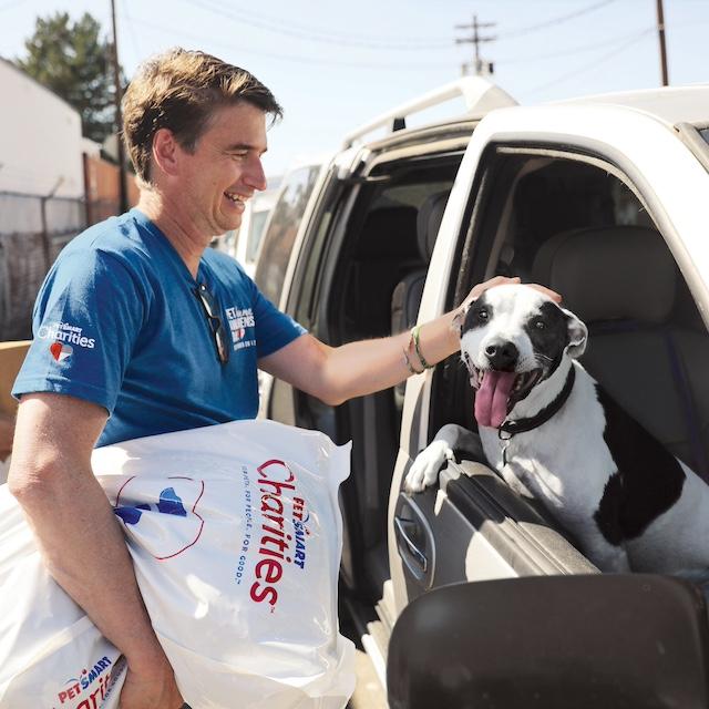 A PetSmart Charities volunteer petting a dog in car and holding a bag of dog food — food insecurity