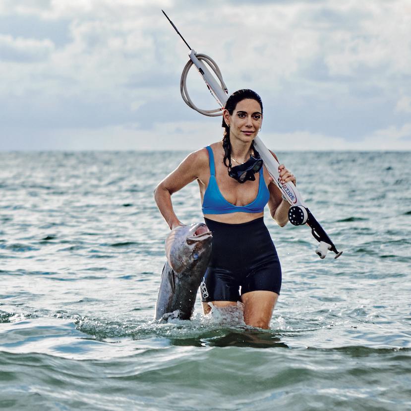 Valentine Thomas, professional spearfisherwoman and sustainable seafood advocate, emerges from the water with a fish and fishing spear.