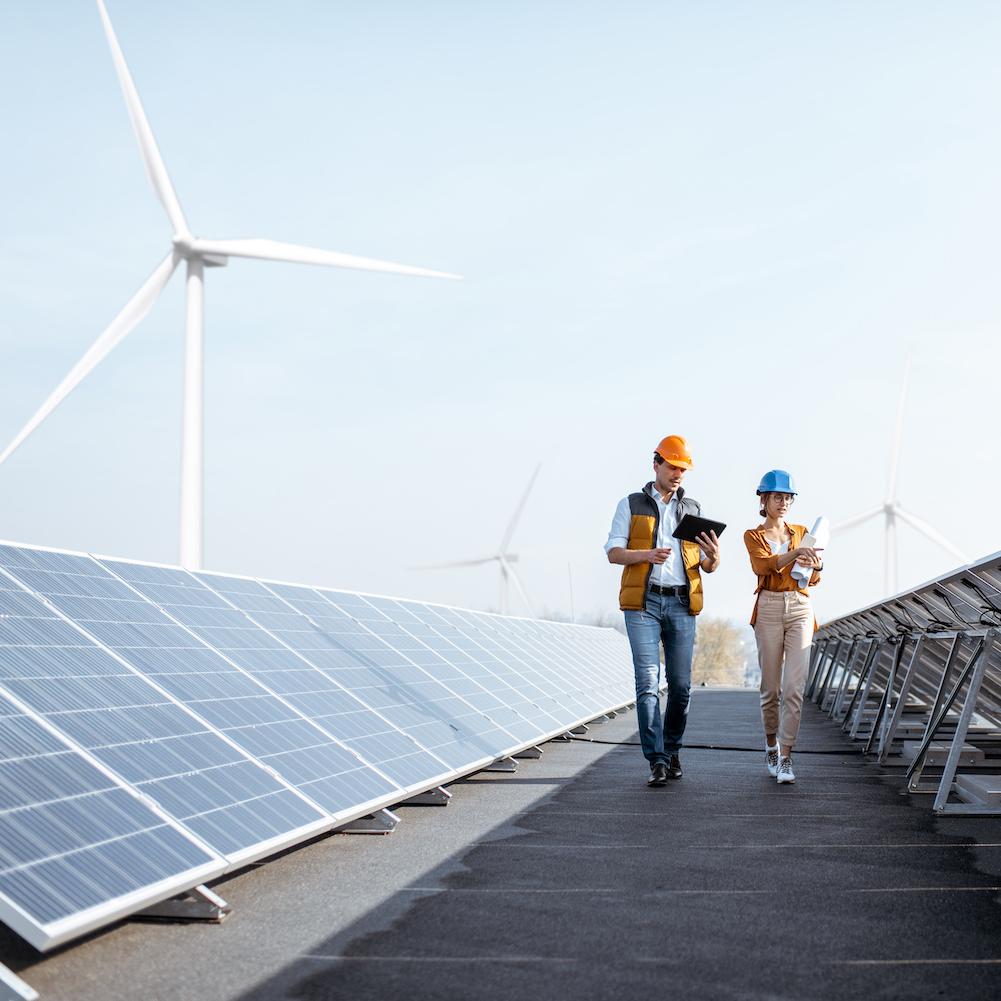 Workers walking through a solar power plant with wind turbines in the background - Achieving the SDGs with renewable energy