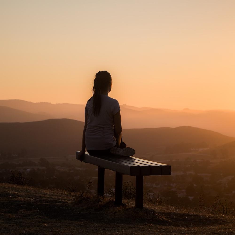 woman sitting meditating on park bench with mountain view at sunset
